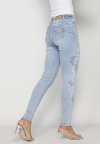 Femme portant un jean skinny bleu clair brodé et des sandales transparentes à talons hauts, posant sur fond blanc.