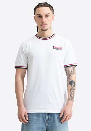 Joven con cabello trenzado que lleva una camiseta blanca de Lonsdale London con ribetes rojos y azul marino, mostrando tatuajes detallados en su brazo derecho.