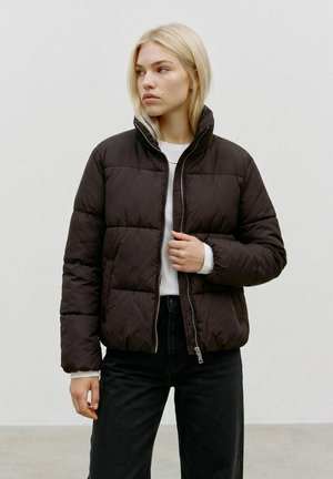 Frau mit blondem Haar trägt eine schwarze Steppjacke über einem weißen Hemd und schwarzen Hosen und steht vor einem schlichten hellgrauen Hintergrund.
