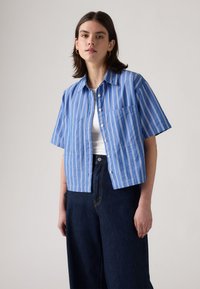 Short-sleeved button-up shirt in light blue with white vertical stripes. Features two front pockets and a classic collar. Worn with dark jeans.
