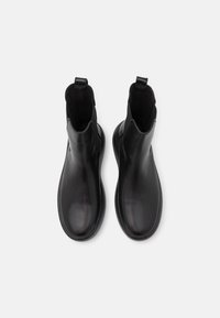 Botines de cuero negro con acabado liso, paneles laterales elásticos y tiradores. Las suelas son planas y ligeramente redondeadas.