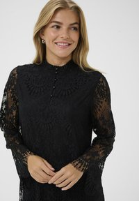 Juoda nėriniuota suknelė su ilgomis rankovėmis, aukštu apykakle ir mygtukų uždarymu. Sudėtingi gėlių raštai visoje suknelėje; permatomos rankovės su banguotais kraštais.