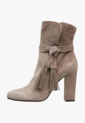 Bottine en daim taupe avec bout pointu, talon carré, et brides décoratives à glands tressés enroulées autour de la tige.