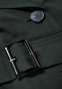 Dark green fabric with a glossy black buckle, silver button detail, and contrasting stitching. Textured surface with button hole.