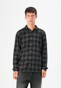 Camisa a cuadros en blanco y negro con diseño de botones, que presenta una textura suave y mangas largas. Modelo lleva jeans oscuros.