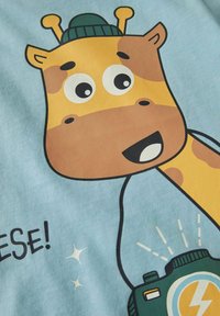 Lysblåt stof med en tegneserie-giraff iført en grøn hat, store øjne og et kamera. Designet inkluderer legende tekst og glitrende stjerner.
