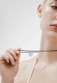 Collar de cuentas negras con una doble cadena, que presenta un colgante redondo de oro con intrincados diseños florales. Fondo en tono de piel.