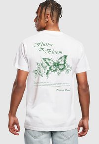 Hvid bomulds t-shirt med en grøn sommerfugl grafik og blomsterdesign på bagsiden, med teksten "Flutter & Bloom" nedenunder.