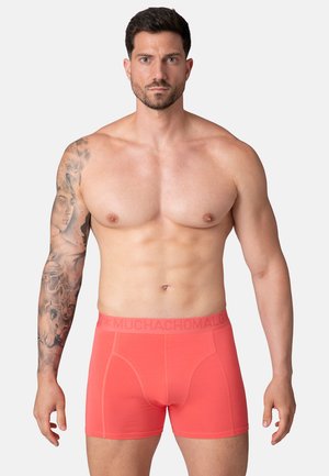 Hombre musculoso con barba y detallado tatuaje en la manga lleva puesto un calzón corto coral, de pie contra un fondo gris claro liso.