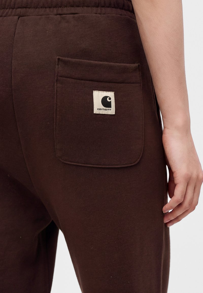 Nahaufnahme einer Person, die braune Hose mit einer quadratischen Taschenklappe auf der Rückseite trägt, welche ein schwarz-weißes Carhartt-Logo zeigt.