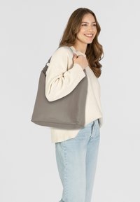 Sac à fourre-tout en cuir gris avec une texture douce, une forme angulaire et une seule sangle, porté sur l'épaule, associé à un pull léger et un jean.