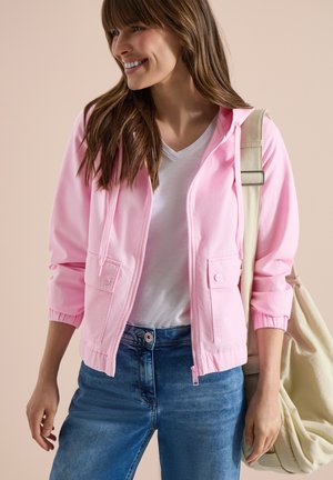 Femme souriante portant une veste zippée rose, un t-shirt blanc en V, un jean bleu et un sac à bandoulière beige.