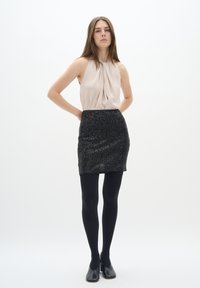 Haut sans manches rose poudré avec un tissu drapé, associé à une mini-jupe noire en sequins. Le mannequin porte des collants noirs et des chaussures à bout fermé.