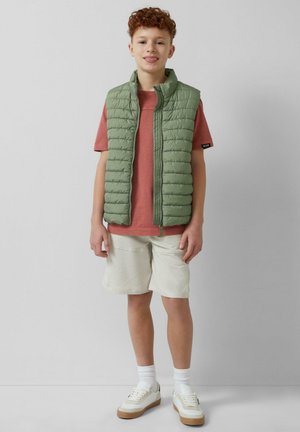 Grüne, wattierte Weste über einem roten Shirt, kombiniert mit hellen Shorts und weißen Sneakern. Die Weste hat horizontalen Nähten und einen Reißverschluss vorne.