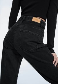 Jeans negros de tiro alto con un ajuste relajado, que presentan bolsillos traseros y una etiqueta de marca de cuero marrón en la cinturilla.
