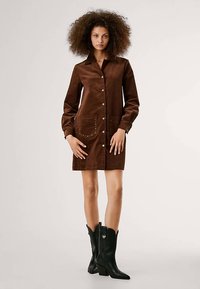Rochie maro din piele întoarsă, cu guler, nasturi în față și două buzunare decorate cu ținte. Modelul o asortează cu ghete negre de gleznă.