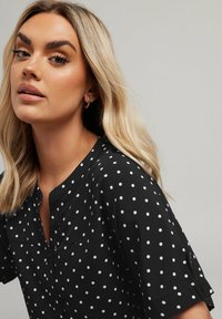 Camicetta nera a pois con vestibilità rilassata, dotata di maniche corte, scollatura a V e motivi circolari bianchi distribuiti in modo uniforme.