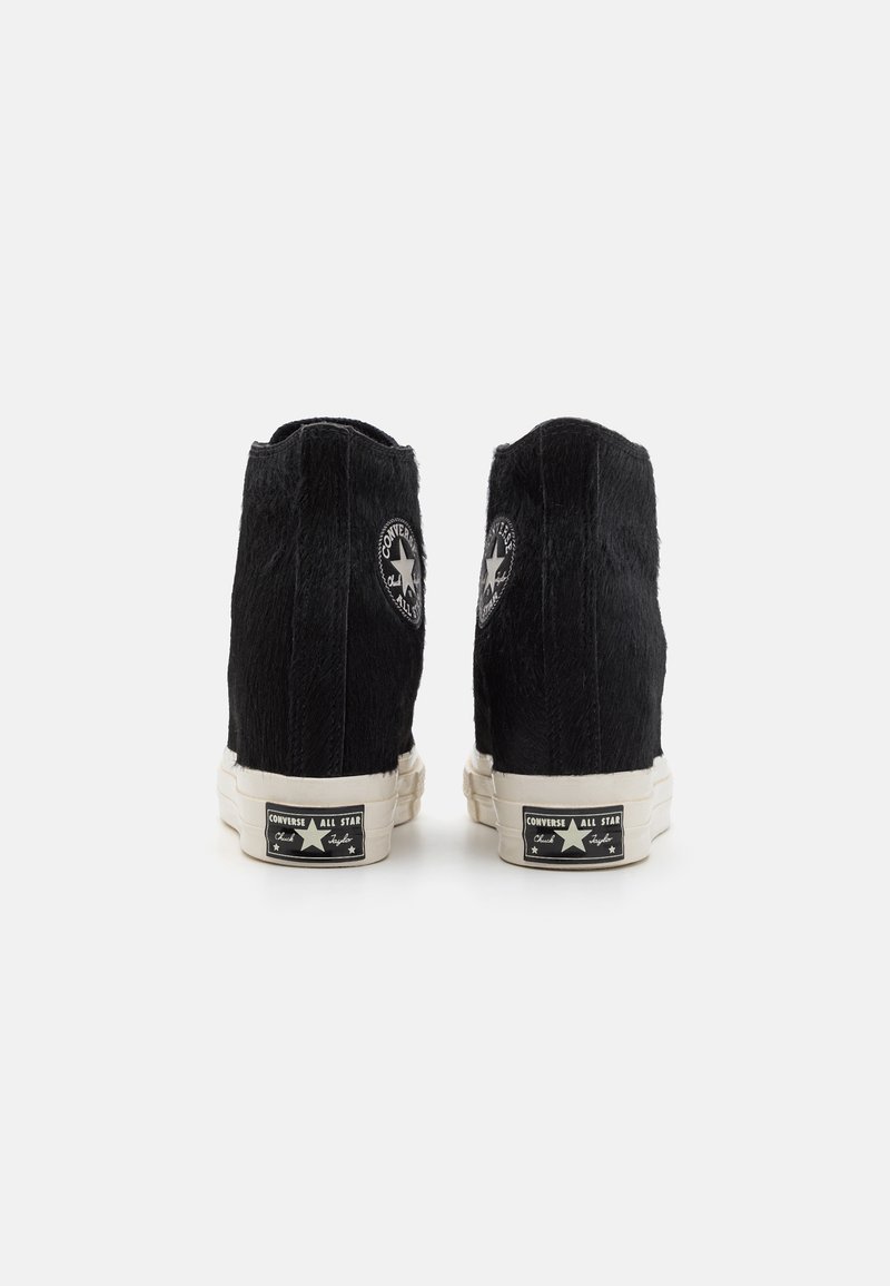 Des baskets montantes noires avec une tige en tissu texturé, une semelle en caoutchouc blanche et des patchs de logo circulaires sur chaque talon. Remarquables pour leur texture unique.