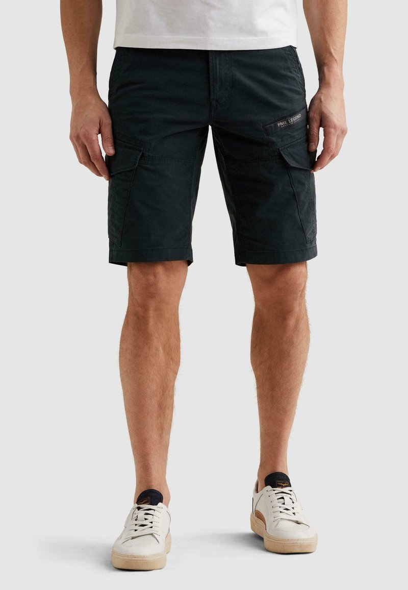 Homme portant un short cargo noir et des baskets blanches, debout devant un fond clair uni.