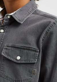 Grå denimskjorta med krage med knappar, två bröstfickor och vita tryckknappar. Har stickade detaljer och en broderad "R"-logotyp.