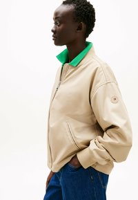 Giacca bomber beige con colletto verde, dotata di zip frontale, polsini a costine, tasche laterali e una toppa con logo sulla manica.