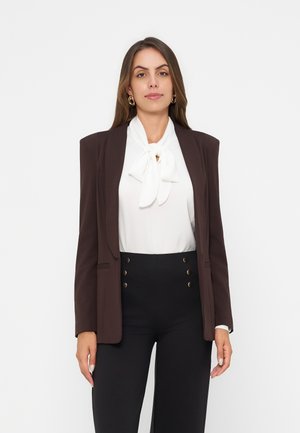 Femme portant un chemisier blanc avec un nœud papillon, un blazer marron foncé avec des épaulettes et un pantalon noir taille haute avec des boutons dorés.