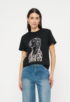 Γυναίκα φοράει μαύρο μπλουζάκι με αφηρημένη γραμμική τέχνη γυναίκας σε φλοράλ τοπ, συνδυασμένο με ψηλόμεσο μπλε τζιν και χρυσούς κρίκους σκουλαρίκια.