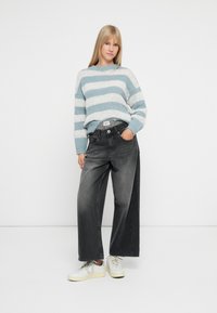 Ung kvinne står med hånden i lomma, iført blå og hvit stripete genser, vide svarte jeans, og hvite sneakers på hvit bakgrunn.