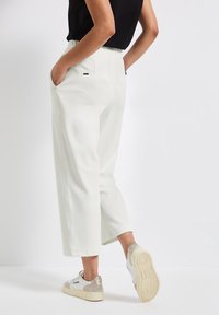 Vita, avslappnade byxor med elastisk midja och sidofickor, i kombination med grå och vita sneakers, sedd bakifrån.