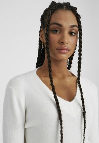 Un pull blanc en tricot doux avec un décolleté en V, présentant des accents côtelés sur le col et une coupe décontractée. Cheveux tressés délicatement coiffés.