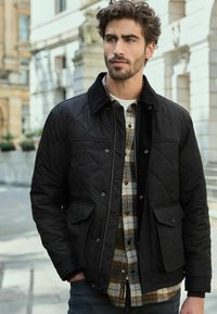 Black quilted jacket with a corduroy collar, two front pockets, worn over a beige and brown plaid shirt, with a white undershirt visible.