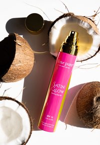 Satin Glow brun utan sol torrolja i en rosa flaska med guldbeslag, omgiven av två bruna kokosnötter, som framhäver SPF 15-skydd.