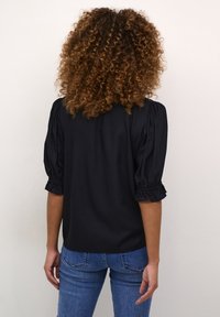 Blouse noire à manches bouffantes, en tissu texturé, avec poignets froncés, associée à un jean bleu ajusté, vue de dos.