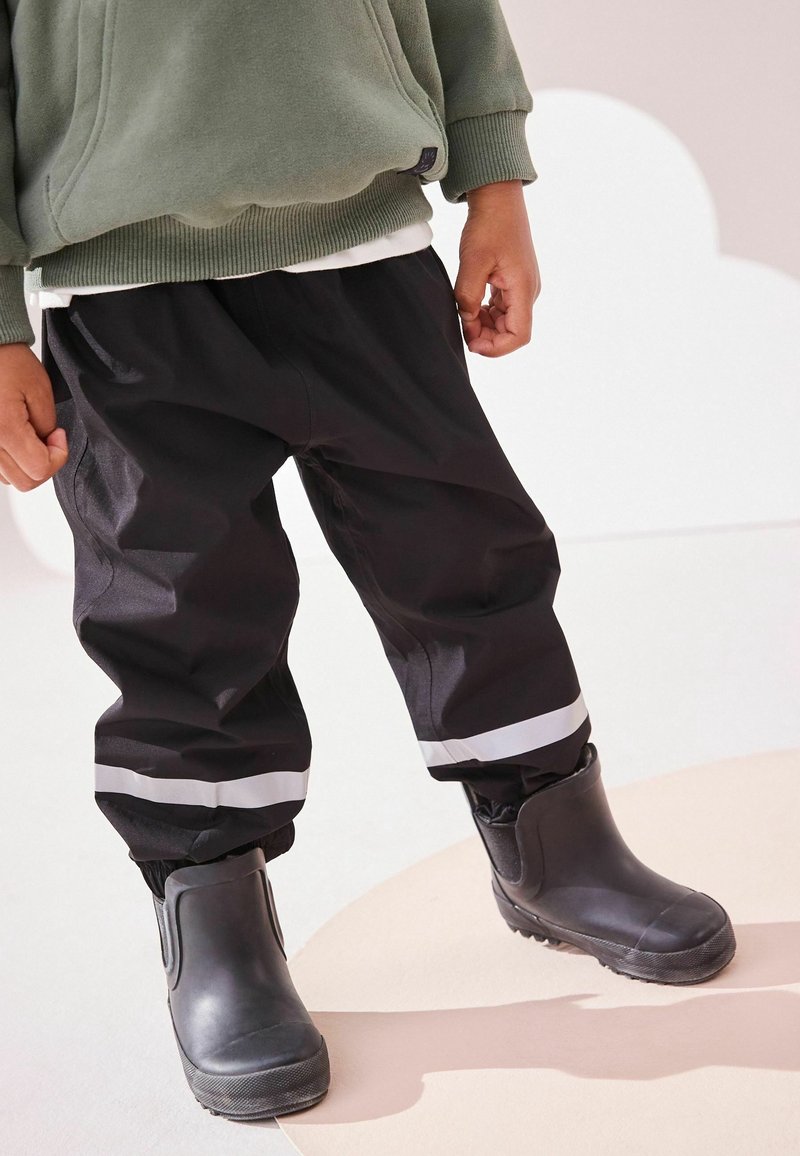 Enfant portant un pantalon imperméable noir avec des bandes réfléchissantes et des bottes en caoutchouc noires, debout sur un sol clair.