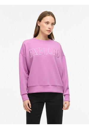 Femme portant un sweat-shirt rose avec le texte "EXPLORE", associée à un pantalon noir, debout devant un fond clair uni.