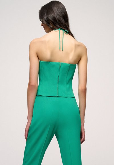 Top verde senza spalline con un design aderente, caratterizzato da una zip sul retro e un dettaglio di legatura al collo. Texture liscia e silhouette sartoriale.