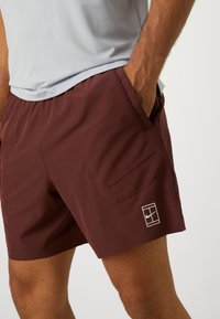 Bordeaux punane spordilühemüts, külgedel taskud, sile tekstuur ja tagasihoidlik Nike logo. Sobitatud heledashalli võrgusärgiga.