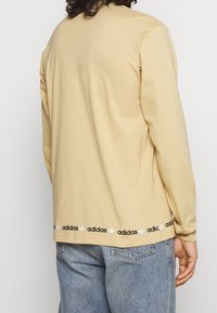 Beige långärmad skjorta med rund halsringning, med ett svart Adidas-logoband vid nederkanten och standardpassform på ärmarna.