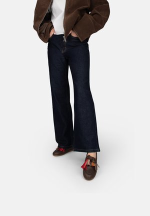 Jean en denim foncé à larges jambes, associé à une veste en daim marron et des chaussures marron ornées de pampilles rouges et multicolores.