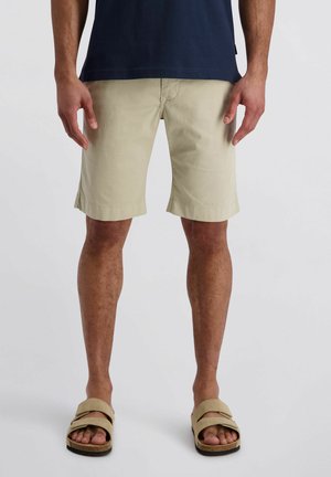 Mann trägt beige kniehohe Shorts, dunkelblaues Hemd und beige Sandalen, steht vor einem schlichten Hintergrund.