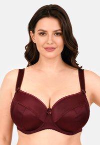 Hullámos barna hajú nő, sötétvörös szatén alátámasztásos melltartót visel, amelyen apró szalagmasnik találhatók a pántokon és a középső előoldalon.
