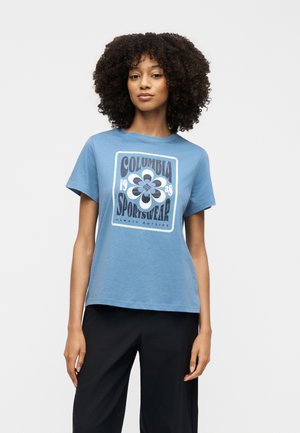 Femme aux cheveux bouclés portant un t-shirt bleu Columbia Sportswear avec un motif de fleur et un pantalon noir, debout devant un fond blanc.