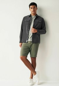 Chemise grise à boutonner avec deux poches poitrine, associée à un short vert olive et des baskets blanches, sur un fond uni.