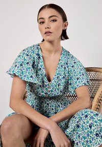 Blumenkleid mit tiefem V-Ausschnitt, kurzen Rüschensleeves und Knopfakzenten. Hergestellt aus leichtem Stoff in Blau- und Grüntönen.