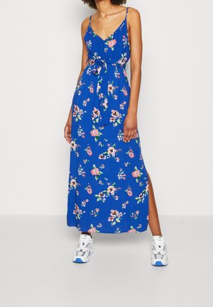 Kvinde iført en blå, blomsterprintet, ærmeløs maxi-kjole med bælte i taljen og hvide sneakers, der står foran en hvid baggrund.