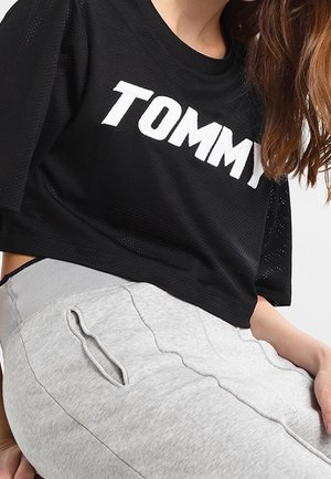 Persona que lleva una camiseta negra con el texto "TOMMY" y pantalones gris claro con un bolsillo lateral, sentada con una mano apoyada en la pierna.