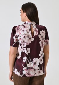 Blusa a maniche corte di un intenso bordeaux scuro con grandi stampe floreali in rosa e bianco, caratterizzata da un dettaglio chiave sul retro e una texture morbida e liscia.