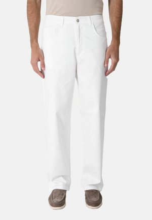 Uomo che indossa pantaloni bianchi a taglio dritto e scarpe marroni slip-on, in piedi davanti a uno sfondo chiaro uniforme.