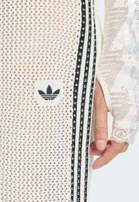 Biely sieťovaný vrch s čiernymi pruhmi a vyšitou čiernou logovou nášivkou. Textúrovaný materiál kontrastuje s hladkými rukávmi zdobenými čipkou.