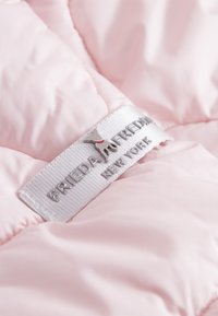 Jasnoróżowy materiał puchowy o gładkiej teksturze. Zawiera srebrną metkę z nazwą marki "FRIEDA KAHLO" oraz małym logo psa.